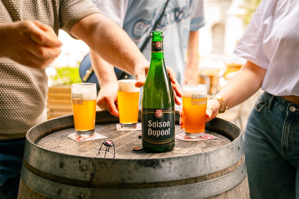 Une bouteille de Saison Dupont et plusieurs verres de Saison Dupont remplis sur un tonneau.
